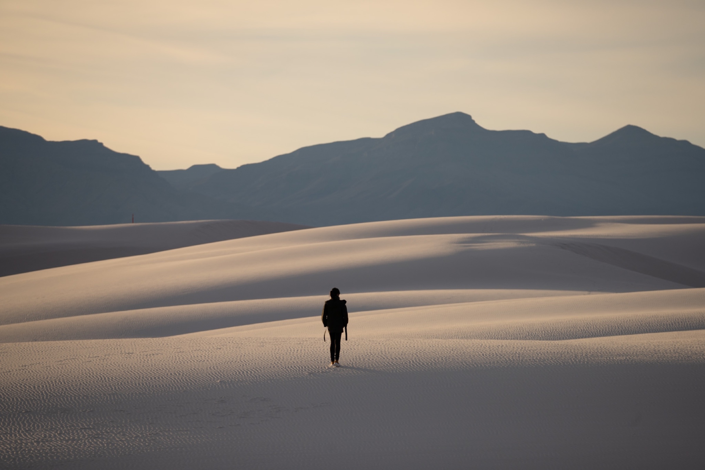 A person walking across white sand dunes with mountains in the distance.